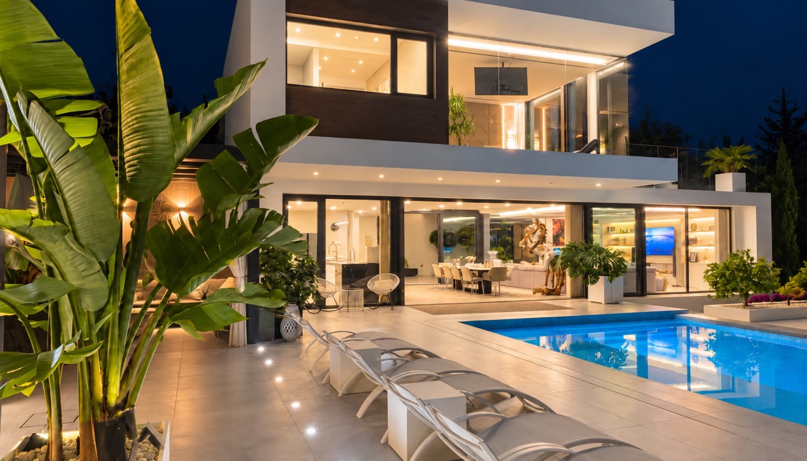 Façade de la villa illuminée la nuit, vue depuis la piscine et les transats